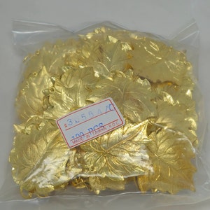 Può includere: Un sacchetto di plastica trasparente pieno di numerosi abbellimenti a forma di foglia metallica color oro. Le foglie hanno venature dettagliate e un piccolo foro per appenderle. Un'etichetta con il numero "30584/0" e "100 PCS" è visibile.
