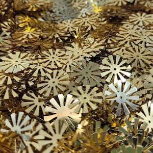 May include: A large collection of small, flat, metallic gold sequins shaped like multi-pointed stars or sunbursts. Each individual sequin features a central circular hole and numerous thin, radiating rays with a reflective, shiny finish.