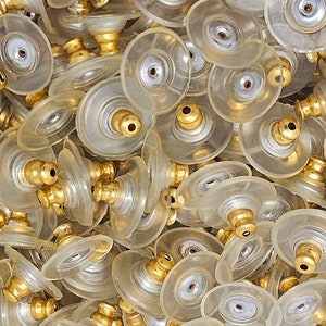 Puede incluir: Primer plano de un montón de cierres de pendientes. Los cierres están compuestos por una pieza de plástico transparente en forma de disco con un pequeño poste de metal dorado. Los postes tienen un pequeño agujero en el extremo. Los cierres de pendientes están dispersos.