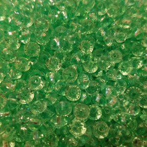 May include: A collection of small, round, translucent green beads with a faceted surface texture. Each bead features a central hole, creating a uniform appearance across the entire group. The light reflects off the angled surfaces of the beads.