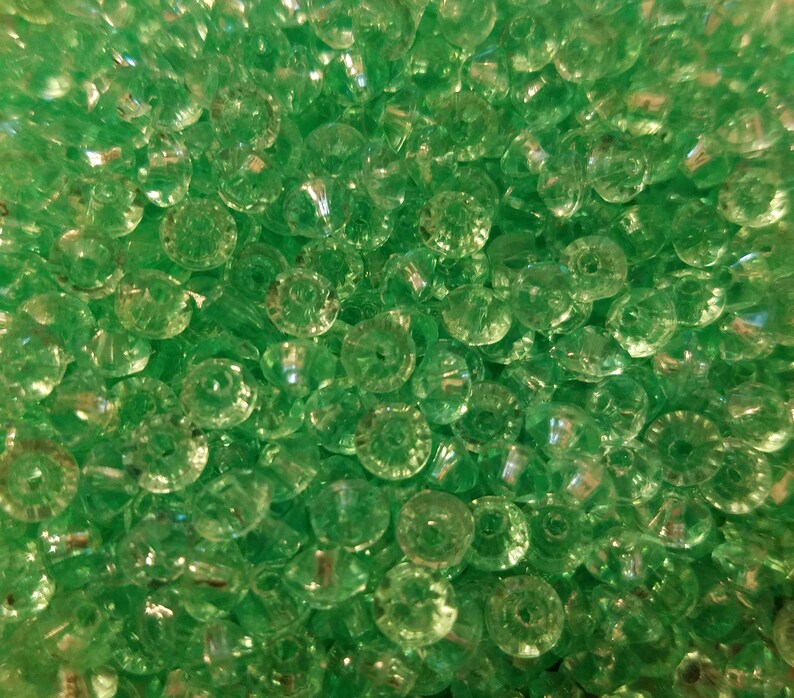 May include: A collection of small, round, translucent green beads with a faceted surface texture. Each bead features a central hole, creating a uniform appearance across the entire group. The light reflects off the angled surfaces of the beads.