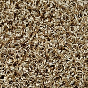 May include: Close-up shot of a pile of gold-colored split key rings. The rings are tightly packed together, creating a dense, textured surface. The rings are made of metal and have a shiny finish.