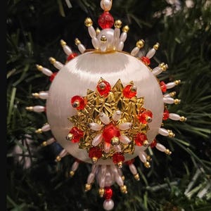 May include: A spherical white satin Christmas ornament featuring a central gold metallic star-shaped embellishment adorned with red faceted beads and white pearls. The perimeter is decorated with radiating white pearl and gold bead spikes.