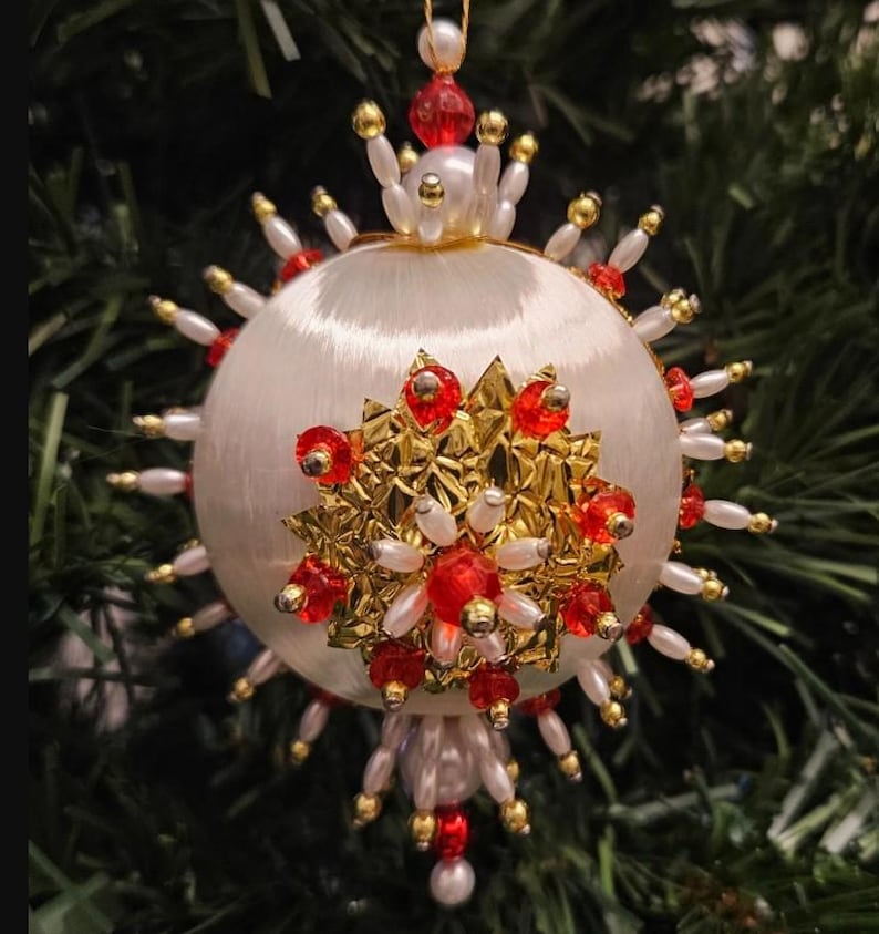May include: A spherical white satin Christmas ornament featuring a central gold metallic star-shaped embellishment adorned with red faceted beads and white pearls. The perimeter is decorated with radiating white pearl and gold bead spikes.