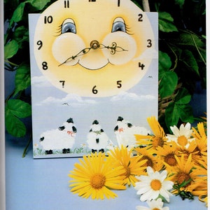 May include: A whimsical, hand-painted clock featuring a smiling sun face with blue eyes and a black, curly mustache. The clock face has black numbers. Below the clock face are three sheep. Yellow and white flowers are in the foreground.