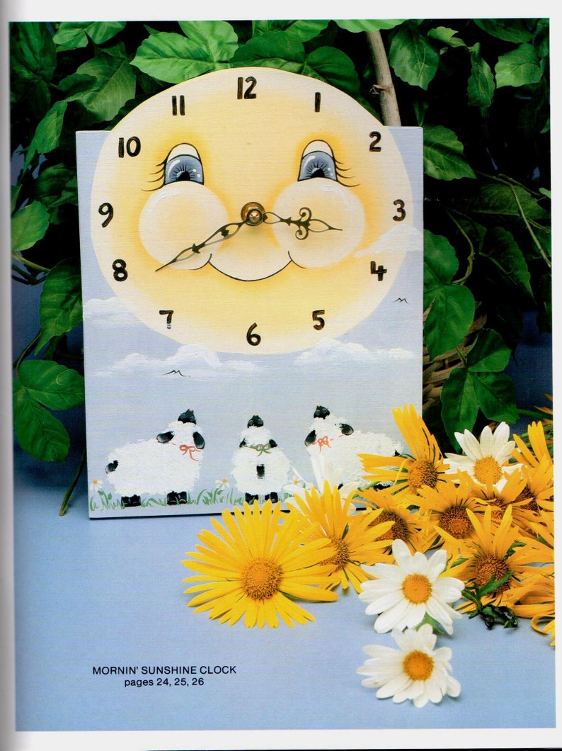 May include: A whimsical, hand-painted clock featuring a smiling sun face with blue eyes and a black, curly mustache. The clock face has black numbers. Below the clock face are three sheep. Yellow and white flowers are in the foreground.