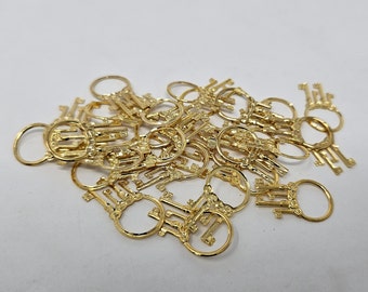 Puede incluir: Una colección de pequeños llaveros dorados. Cada llavero presenta un diseño de llave con un anillo circular para la fijación. Los llaveros están dispersos sobre una superficie blanca, creando una disposición visualmente atractiva.
