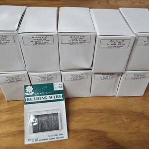 Peut inclure: Plusieurs boîtes blanches et une carte de fil à perler sont présentées. Les boîtes sont étiquetées avec des détails tels que "Beading Wire" et "34 Gauge Silver". La carte affiche "Superior Quality Beading Wire" et le fil. Les articles sont sur une surface en bois.