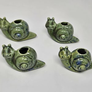 May include: Four ceramic snail ornaments in shades of green with blue detailing. Each snail has a round body with a hole on top, and a smiling face. The ornaments are on a white surface.
