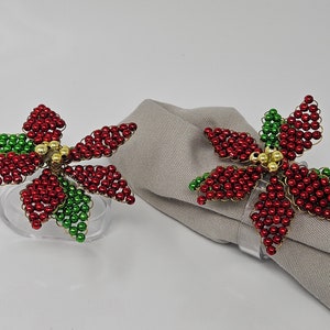 May include: Two red and green beaded poinsettia napkin rings. The rings are made of small red and green beads, with a gold center. The rings are on a gray napkin.
