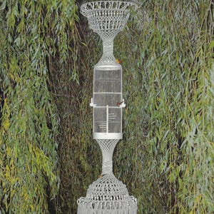 May include: A tall, ornate macrame birdcage structure with a white metal cage in the center. The structure has a flared top and base, with a fringe detail. The text "Macrame Enchantment" and "Paradise" are visible.