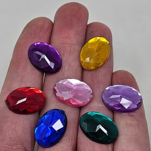 Peut inclure: Sept strass ovales de différentes couleurs, dont le violet, le rouge, le rose, le jaune et le vert. Les strass sont disposés sur une main.