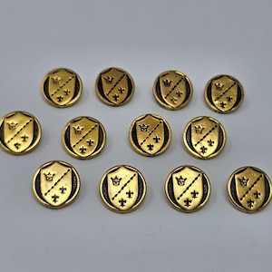 Puede incluir: Doce botones de metal de color dorado con un diseño de escudo y una corona en cada escudo. Los botones tienen un fondo negro y detalles dorados.
