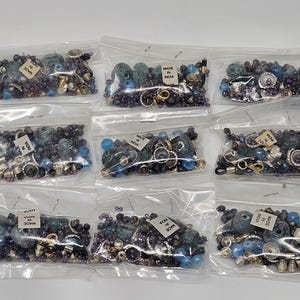 May include: Nine clear plastic bags filled with an assortment of blue, teal, and silver-toned beads, charms, and metal findings. Each bag contains a small white label printed with the text Made in India, showcasing a variety of textures and shapes.