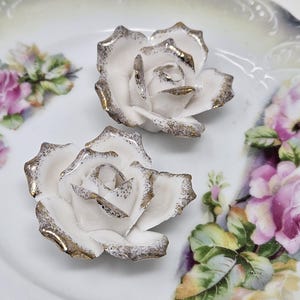 May include: Two white porcelain rose figurines with gold trim. The roses are set on a decorative plate with a floral design in pink, green, and yellow. The plate has a scalloped edge.