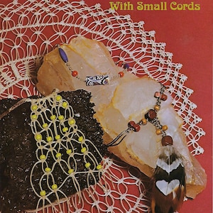 May include: A book cover with the title "How to Macrame' With Small Cords". The cover features a red background with a white crocheted doily and two macrame necklaces on top of a white rock.