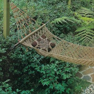 Puede incluir: Una hamaca de macramé beige con cojines marrones cuelga en un exuberante jardín verde. La hamaca está hecha de cuerda y cuentas de madera. El texto "Swing Easy Hammock - Page 18" y "Table Cover - Page 22" es visible.