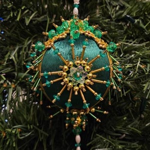 May include: A spherical Christmas ornament featuring a dark green satin fabric base. The surface is intricately decorated with gold-toned metallic beads, clear green faceted beads, and small white pearls arranged in a starburst pattern.