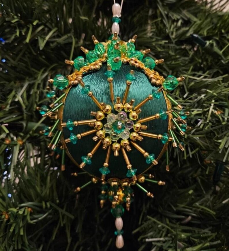 May include: A spherical Christmas ornament featuring a dark green satin fabric base. The surface is intricately decorated with gold-toned metallic beads, clear green faceted beads, and small white pearls arranged in a starburst pattern.