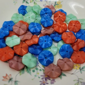 May include: A collection of colorful flower-shaped buttons in shades of blue, green, orange, and brown. The buttons are made of glass and have a slightly iridescent finish.