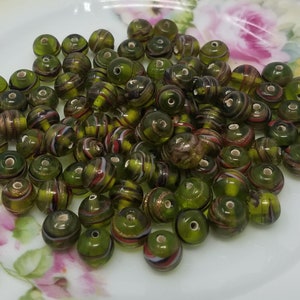 May include: A pile of green glass beads with gold, red, and white stripes. The beads are round and have a hole in the center.