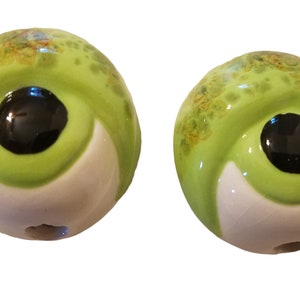 May include: Two green ceramic beads with white and black accents, resembling eyes. The beads have a glossy finish and a small hole for stringing.