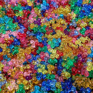 May include: A close-up image of a large pile of colourful plastic beads. The beads are small and round, and they are arranged in a random pattern. The colours of the beads include red, orange, yellow, green, blue, purple, and pink.
