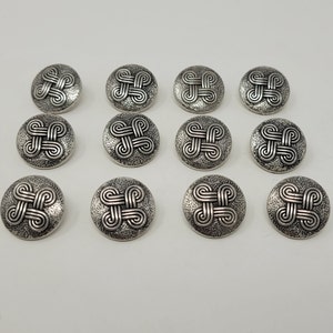Puede incluir: Doce botones plateados con un diseño decorativo e intrincado. Cada botón presenta un patrón repetitivo de líneas entrelazadas.
