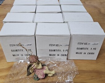 Puede incluir: Una colección de cajas de cartón blancas con texto impreso, cada una con una pieza. Una pequeña figura de ángel en rosa y verde está frente a las cajas. El ángel está acostado y tocando una flauta. Las cajas están etiquetadas como "ITEM NO." y "MADE IN CHINA."