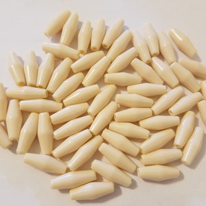 Puede incluir: Un montón de cuentas ovaladas lisas de color blanco roto. Las cuentas están hechas de un material liso y duro y tienen un acabado ligeramente brillante.