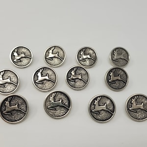 Puede incluir: Doce botones de metal plateados con un diseño de ciervo. Cada botón es redondo y tiene una silueta de ciervo en relieve.