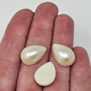 Pode incluir: Três cabochões de resina em forma de lágrima de cor branca perolada. Dois dos cabochões têm um acabamento brilhante, enquanto o terceiro tem um acabamento fosco.