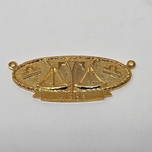 May include: Gold-colored metal charm with a Libra zodiac symbol and the word "LIBRA" inscribed on a banner.