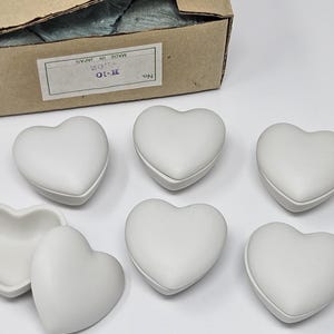 Puede incluir: Una colección de joyeros blancos en forma de corazón, algunos con tapas, dispuestos sobre una superficie blanca. Una caja de cartón es visible en el fondo. Es probable que las cajas estén hechas de cerámica o un material similar.