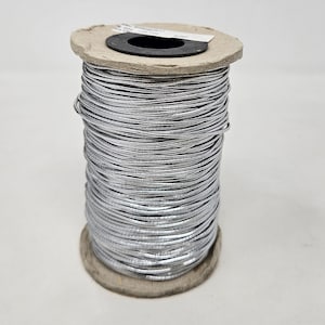 May include: A spool of silver metallic cord wrapped around a cardboard spool. The cord is wound tightly and has a shiny, reflective finish.