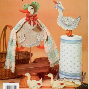 Puede incluir: Un conjunto de figuritas de ganso de madera, pintadas en un estilo caprichoso. La ganso m&aacute;s grande lleva un sombrero y una bufanda, y los gansos m&aacute;s peque&ntilde;os sostienen corazones. El texto "Gwendolyn Goose" y "Gilda Goose" est&aacute; impreso en la parte superior de la imagen. El texto "Geese on the Go" est&aacute; impreso en la parte inferior de la imagen.