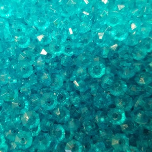 May include: A large collection of small, translucent turquoise blue beads with a faceted, multi-sided cut. Each bead features a central hole and a reflective, crystalline surface that catches the light, creating a dense, uniform texture.