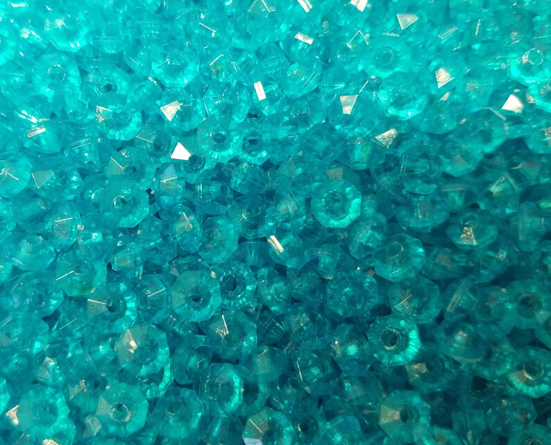 May include: A large collection of small, translucent turquoise blue beads with a faceted, multi-sided cut. Each bead features a central hole and a reflective, crystalline surface that catches the light, creating a dense, uniform texture.