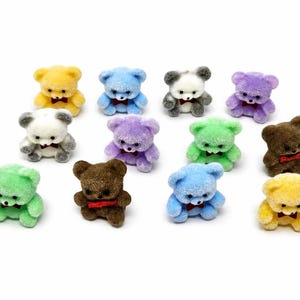 May include: A collection of twelve miniature flocked teddy bear figurines in various colors including yellow, blue, white, purple, green, and brown. Each small bear features a textured fuzzy surface, black eyes, and a tiny red bow tie at the neck.