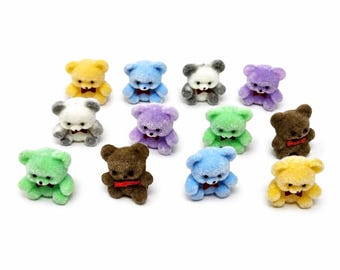 Puede incluir: Una colección de doce figuras en miniatura de osos de peluche flocados en varios colores, incluyendo amarillo, azul, blanco, morado, verde y marrón. Cada pequeño oso presenta una superficie texturizada y difusa, ojos negros y una pequeña pajarita roja.