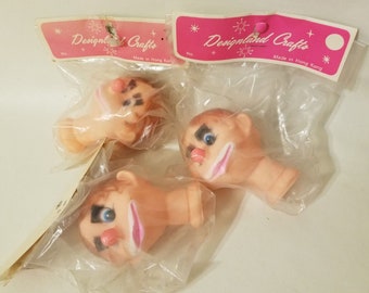 May include: Three plastic doll heads with painted facial features, including eyebrows, eyes, noses, and mouths. The heads are packaged in clear plastic bags with pink and white labels that read "Designland Crafts" and "Made in Hong Kong".