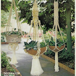 May include: Three macrame plant hangers with white cord and tassels. The hangers are hanging from a wooden beam. The hangers are labeled "Decorator Delight", "Hanging Garden", and "Touch of Glamour".