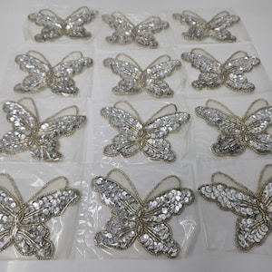 May include: Twelve silver and gold butterfly appliques on white packaging. The butterflies are made of sequins and have a delicate, feminine design.