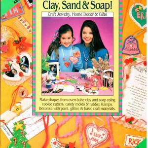 May include: A book cover with the title "Kids Can Do It, Too! Fun with Clay, Sand & Soap!"  The cover features two girls making crafts with clay, sand, and soap. The book is about craft jewelry, home decor, and gifts.