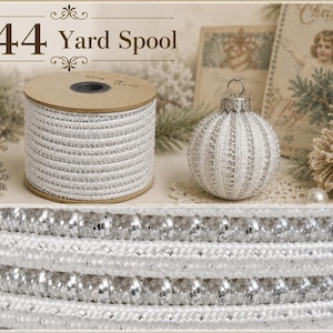 Könnte beinhalten: Eine 144-Yard-Spule mit silbernem und weißem Metallic-Kordelband in texturiertem, geflochtenem Design. Die Spule wird neben einem passenden silber-weißen Deko-Ornament präsentiert, das das komplizierte, schimmernde Gewebe der Kordel zeigt.