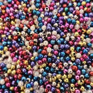 Puede incluir: Primer plano de un montón de pequeñas cuentas de colores. Las cuentas son una mezcla de colores, incluyendo rojo, azul, morado, dorado y blanco. Son redondas y tienen un pequeño agujero en el centro. Las cuentas probablemente se utilizan para hacer joyas u otras manualidades.