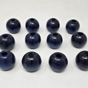 May include: Twelve round, dark blue wooden beads with a hole through the center. The beads are evenly spaced on a white surface. The beads have a smooth, polished finish and are likely for crafting or jewelry making.