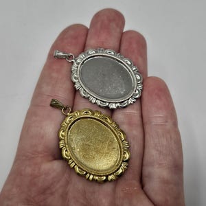Puede incluir: Dos colgantes de metal de forma ovalada con un marco decorativo de borde festoneado. Un colgante tiene un acabado en tono plateado brillante, mientras que el otro tiene un tono dorado. Cada colgante incluye una pequeña anilla metálica superior.