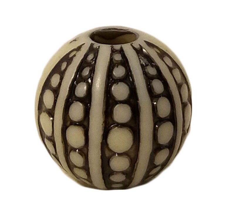 May include: A white bead with a brown and white striped pattern. The bead has a round shape and a smooth surface.