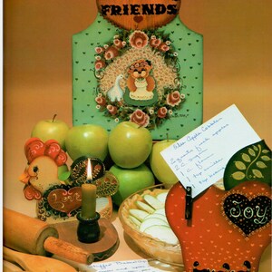 Puede incluir: Un letrero de madera pintado con las palabras "Welcome Friends" en forma de coraz&oacute;n. El letrero es verde con un patr&oacute;n de lunares blancos y una corona de flores con un oso de peluche en el centro. Hay manzanas, una vela, un rodillo y una gallina de madera con la palabra "Love" pintada en ella. Tambi&eacute;n hay una manzana roja con la palabra "Joy" pintada en ella. El texto "Chicken Love" y "Apple of Joy" est&aacute; escrito en la imagen.